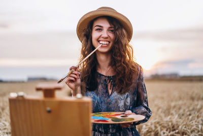 A young woman with curly hair and wearing a hat is painting in nature. 