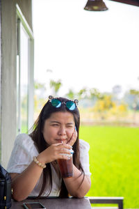 An asian woman in a white dress is sucking a soft drink on a table with a rice field background.