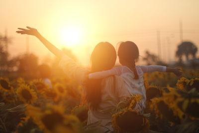 Rear view of mother and daughter with arms outstretched at sunflower field during sunset