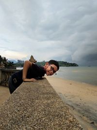 Portrait of boy leaning on retaining wall at beach against cloudy sky