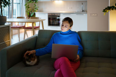 Woman freelancer sits on sofa in living room petting loving cat and watching webinar in laptop