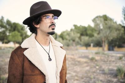 Handsome young bearded male in eyeglasses wearing trendy warm coat and hat looking away thoughtfully while standing against blurred trees in autumn day