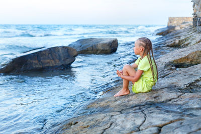 A little girl is sitting on a rocks by the sea. long braided hair. gaze into a distance. 