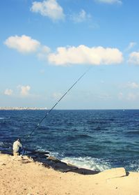 Man with fishing rod sitting at sea shore against sky
