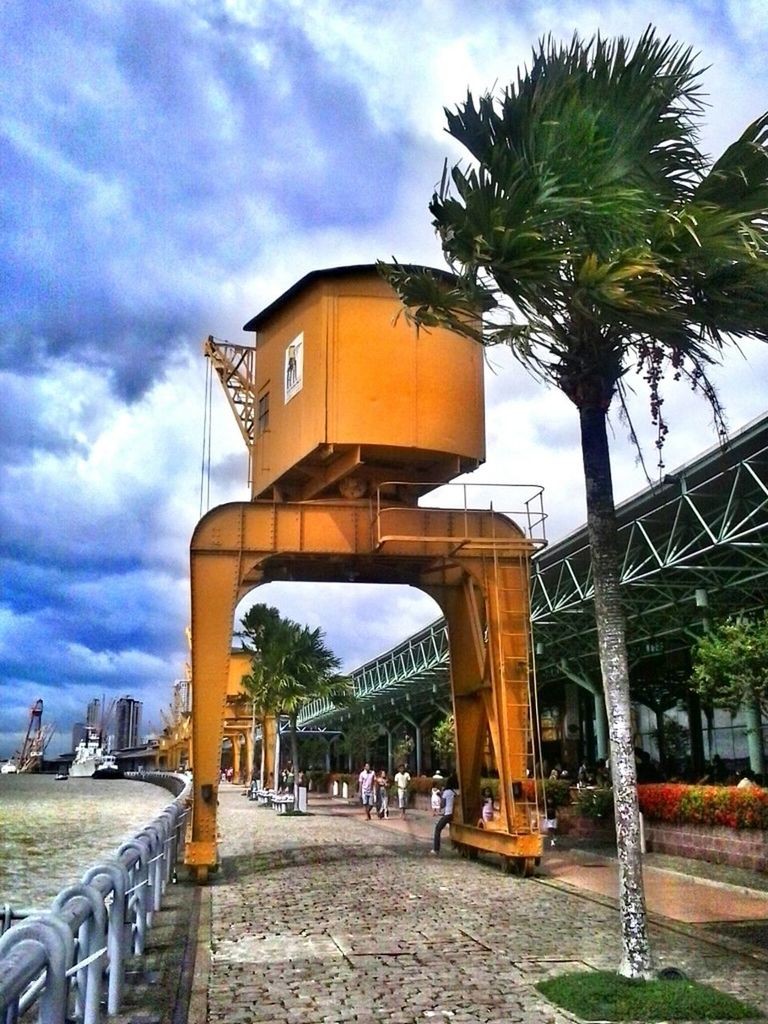 sky, tree, built structure, architecture, cloud - sky, cloud, building exterior, palm tree, cloudy, incidental people, outdoors, day, railing, nature, growth, transportation, footpath, sunlight, beach, water