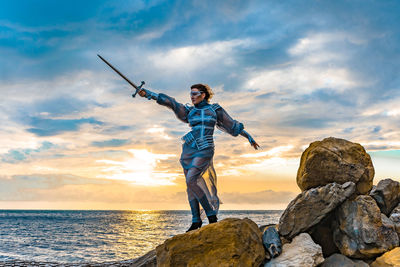 Rear view of man with arms raised standing on rock against sky during sunset