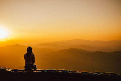 Silhouette man sitting on mountain against orange sky