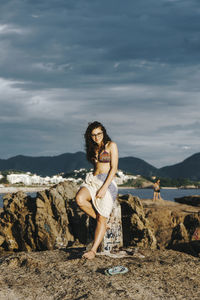 Portrait of young woman standing at beach against sky