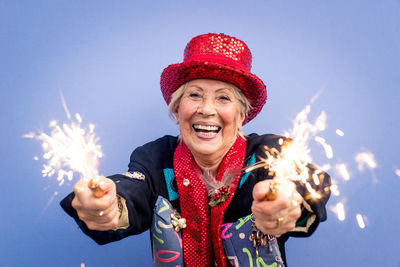 Portrait of happy senior woman wearing hat holding sparklers against blue background