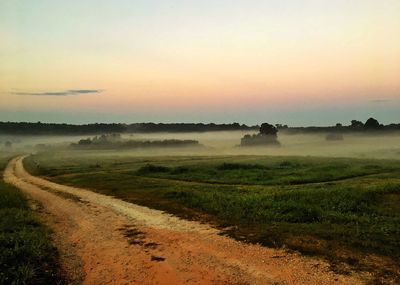 Dirt road amidst field against sky during sunset