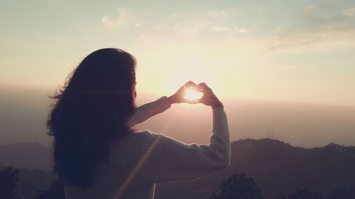 Woman making heart shape from hands in front of sun against sky