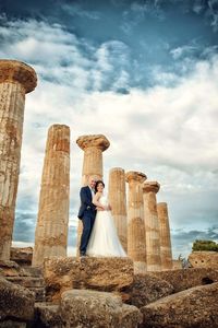 Low angle view of bride and groom by old ruin columns standing against cloudy sky