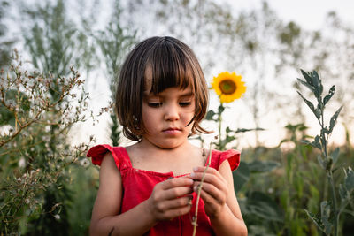 Thoughtful girl in red dress watching small sunflower outside