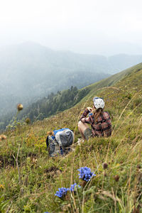 Young woman sitting on meadow in mountain valley next to backpack drinking water  on hike landscape 