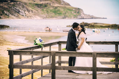 Couple standing on retaining wall by sea against sky