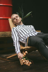 Portrait of young man holding flowers while relaxing on chair