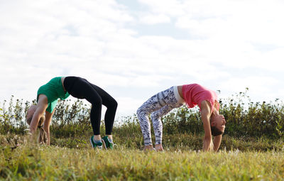 Full length of friends doing yoga on grassy field against sky