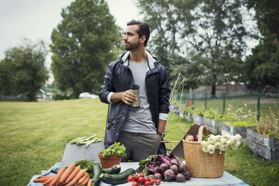 Mid adult man having coffee while standing by freshly harvested vegetables on table at urban garden