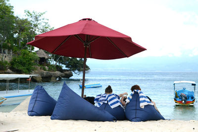 Man and woman relaxing on cushion under umbrella at beach against sky
