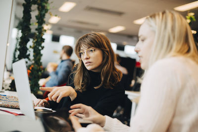 Young businesswoman pointing at laptop to female colleague sitting in office