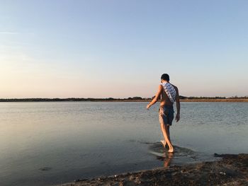 Rear view of shirtless man walking at lakeshore during sunset