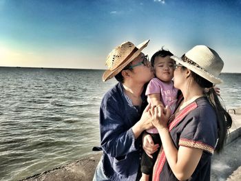 Parents kissing daughter while standing by sea against sky