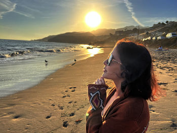 Side view of young woman standing at beach against sky during sunset