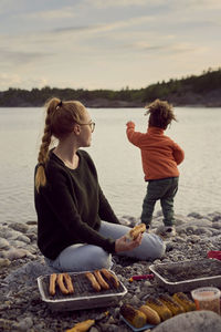 Mother sitting by barbecue grill while looking at dancing son at beach against sky