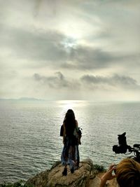 Rear view of woman standing on rocks at beach against cloudy sky