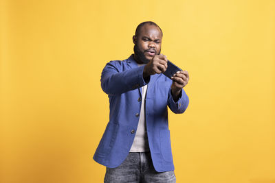 Portrait of young man using mobile phone while standing against yellow background