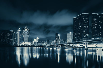 Illuminated modern buildings by river against sky at night