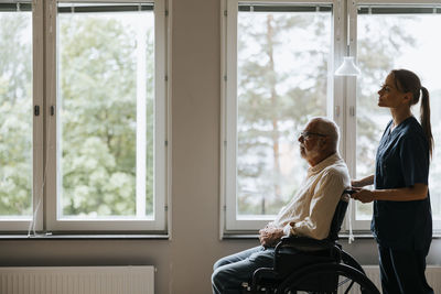 Female nurse helping senior man sitting in wheelchair near window at nursing home