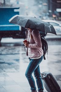 Woman carrying umbrella while walking with wheeled luggage during rainy season