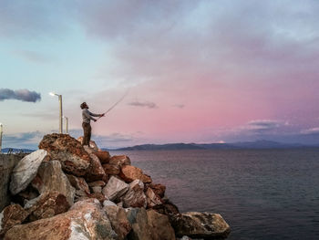 Fisherman on rock formation by sea against sky during sunset