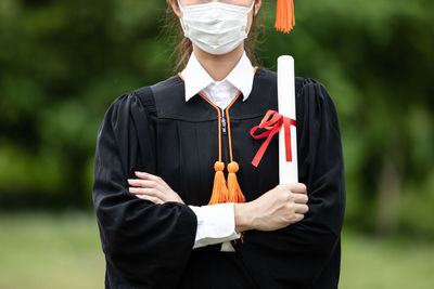 Midsection of student wearing mask holding diploma in hand standing outdoors