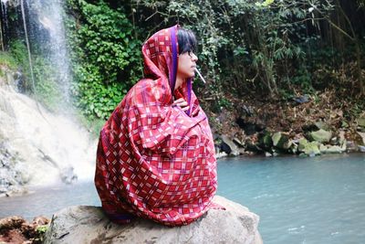 Young man wrapping in blanket sitting on rock by river