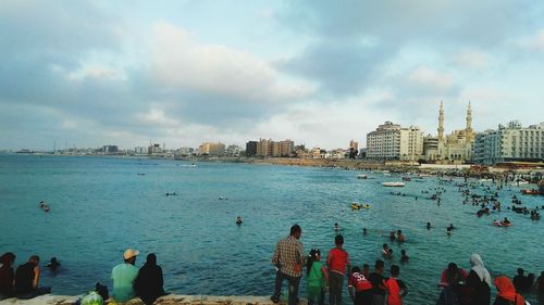 People on sea by cityscape against sky