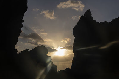 Low angle view of sunlight streaming through silhouette rocks against sky
