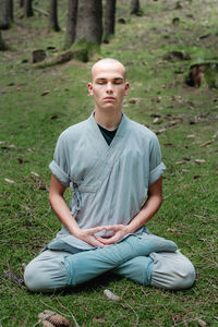 Bald man in traditional clothes sitting on grass in lotus pose and meditating during kung fu training in forest