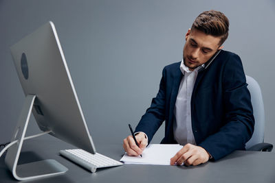 Young businessman writing on paper while talking on mobile phone at desk