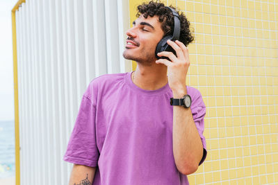 Glad hispanic man in purple t shirt adjusting wireless headphones and smiling with eyes closed while listening to favorite music near yellow tiled wall on weekend day on beach