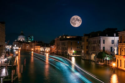 Light trails on river amidst buildings against sky at night