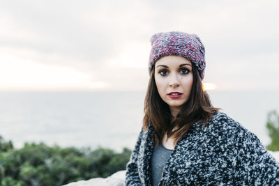Portrait of young woman wearing sweater and knit hat while standing against sea