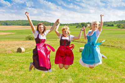 Smiling female friends with dirndl jumping over field against sky