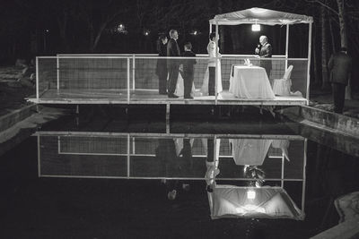 People standing with bride and bridegroom over pond on footbridge at night