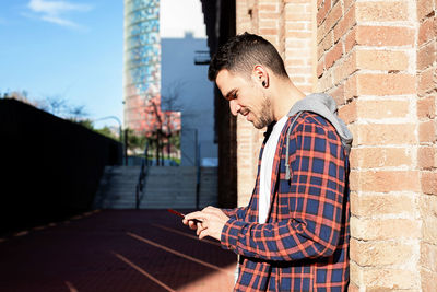 Young man using phone while standing against wall