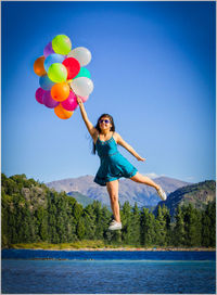 Full length of smiling young woman with balloons against blue sky