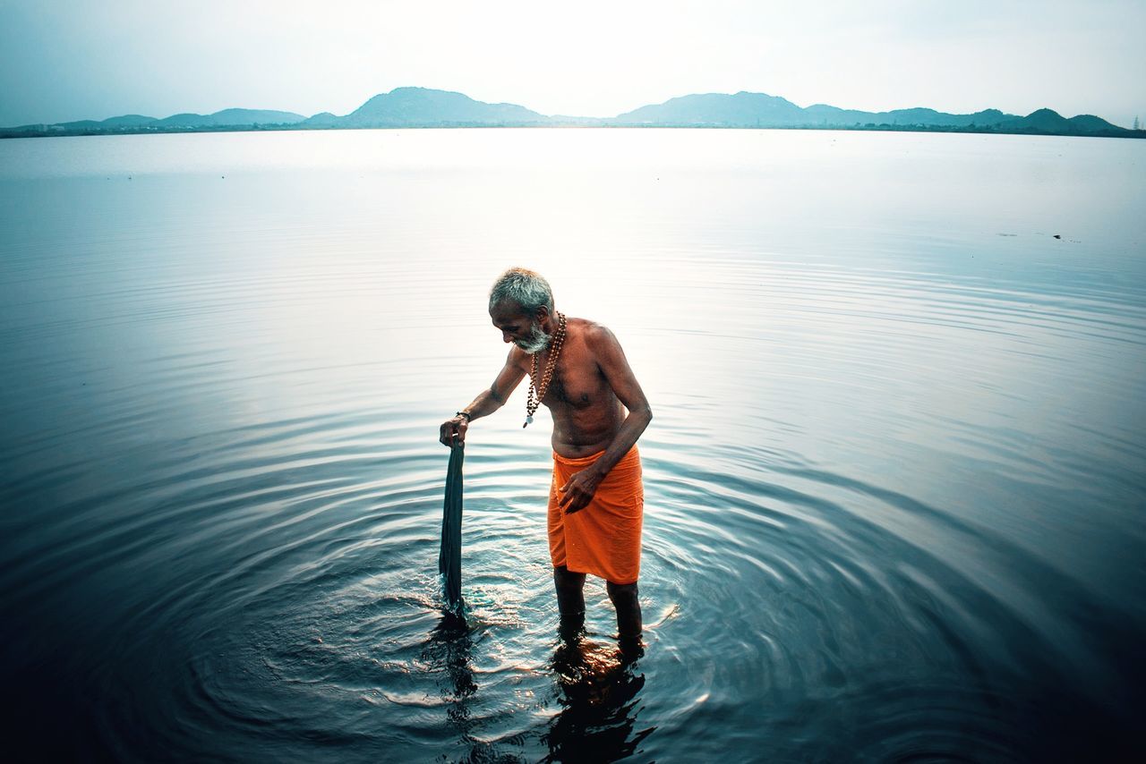 water, one person, reflection, nature, sea, sky, adult, men, beauty in nature, standing, mountain, tranquility, scenics - nature, full length, outdoors, rippled, tranquil scene, activity, ocean, occupation, day, environment, clothing, sunlight, fishing, lifestyles, wave, solitude, holiday, land, travel, beach