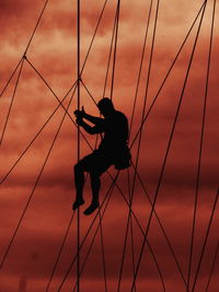 Low angle view of silhouette worker climbing on cable against cloudy sky during sunset