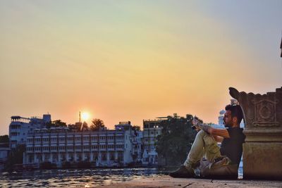 Side view of man leaning on retaining wall by river in city during sunset